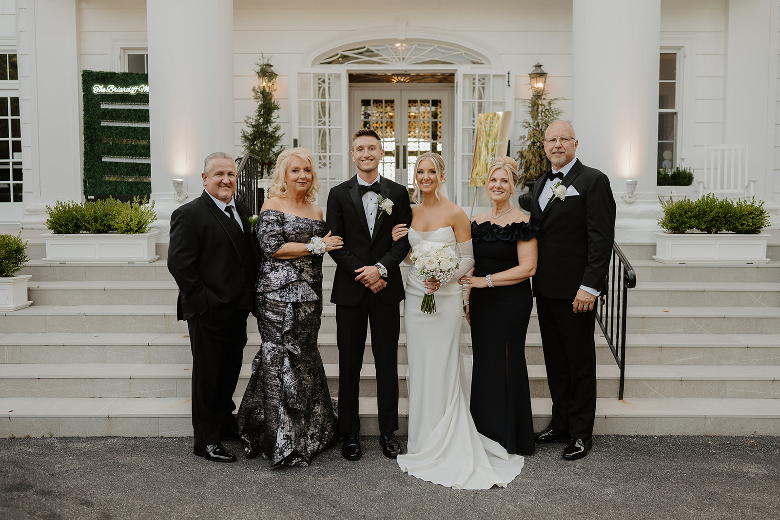 Newlyweds pose with their parents for a formal group portrait on the steps of the white manor.